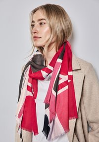 Veelkleurige sjaal in rood, zwart en roomkleur met abstracte patronen en franjes aan de randen, gedrapeerd om de hals over een lichte top.