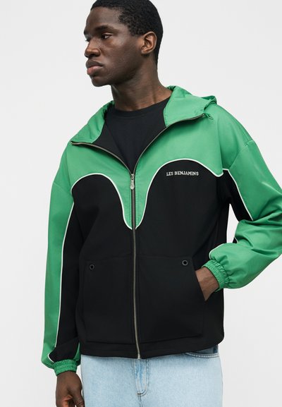 Νεαρός άνδρας που φοράει πράσινο και μαύρο μπουφάν με φερμουάρ και το κείμενο «LES BENJAMINS» στο στήθος, συνδυασμένο με ανοιχτόχρωμα μπλε τζιν, στέκεται μπροστά σε λευκό φόντο.