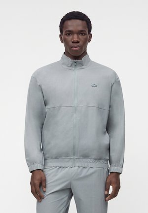Jeune homme portant une veste zippée gris clair à col montant et un pantalon assorti, debout devant un fond blanc uni.