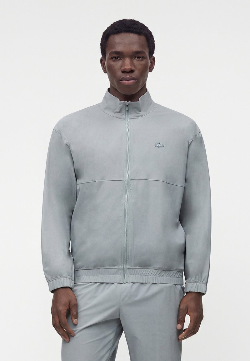 Jeune homme portant une veste zippée gris clair à col montant et un pantalon assorti, debout devant un fond blanc uni.
