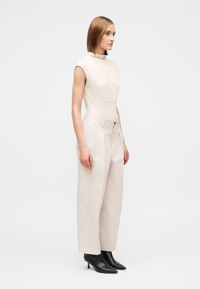 Beige vartaloa myötäilevä body korkealla kauluksella, yhdistettynä löysiin kermaan vivahtaviin housuihin ja mustiin terävälestisiin nilkkureihin. Minimalistinen muotoilu, sileä tekstuuri.