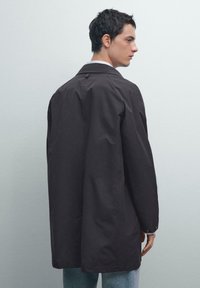 Cappotto lungo marrone scuro realizzato in tessuto liscio. Presenta un ampio colletto, polsini con bottoni e una linea dritta. Indossato sopra jeans blu chiaro.