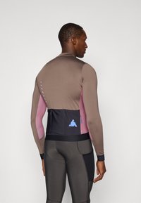 Langærmet rash guard i brun og pink stof, med netryg, logo detaljer og tætsiddende design. Bæres med mørke leggings.