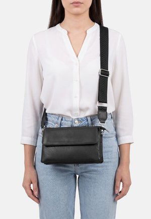 Donna che indossa una camicia bianca con bottoni e jeans azzurri chiari con una borsa nera in pelle a tracolla con a tracolla larga regolabile.
