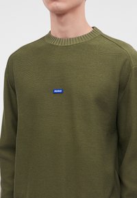 Homme portant un pull en maille vert olive avec col rond côtelé et petit écusson logo bleu "HUGO" centré sur la poitrine.