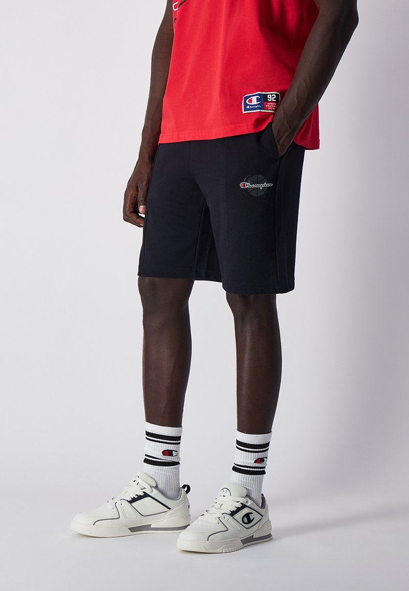 Pantalones cortos de algodón negros con bolsillos laterales, con un logo de Champion en la pierna izquierda, combinados con una camiseta roja y zapatillas blancas.