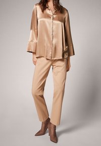 Camicia lunga in satin beige con colletto abbottonato, abbinata a pantaloni beige tagliati su misura e tacchi a punta marroni, con una texture liscia.