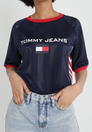 Navy blue short-sleeved shirt with red accents and stripes, featuring "TOMMY JEANS" logo and a small rectangular flag design. Worn with light denim jeans.