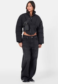 Femme portant une veste matelassée noire courte, un jean large noir, des baskets noires et un sac bandoulière noir avec une pochette fixée.