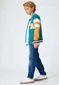 Chaqueta de béisbol en color teal y naranja con rayas blancas, que presenta un gran parche de "LA", combinada con jeans azules y zapatillas blancas.