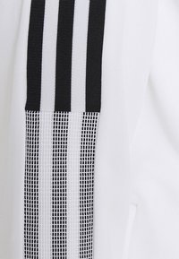 Vit träningsjacka med tre vertikala svarta ränder, en med ett texturerat nätmönster, längs vänster sida för kontrast och stil.