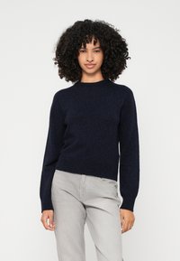 Mornarji modra pletena jopica s krožnim izrezom, dolgimi rokavi in ribastim robom. Ujemanje s svetlo sivo hlačami, minimalistična zasnova.