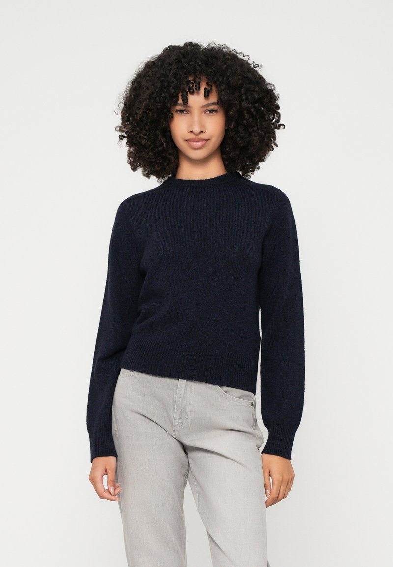 Mornarji modra pletena jopica s krožnim izrezom, dolgimi rokavi in ribastim robom. Ujemanje s svetlo sivo hlačami, minimalistična zasnova.