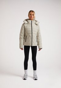 Lys grå quiltet jakke med høj krave og aftagelig hætte, med sort lynlås og detaljer. Bæres med sorte leggings og sneakers.