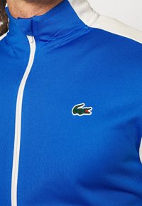 Blå zip-jacket med hög krage och vita accenter. Har textil med struktur och en grön krokodil-logotyp broderad på bröstet.