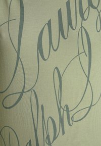 Tissu vert clair avec des inscriptions cursives tourbillonnantes en vert foncé. La texture semble lisse avec un léger éclat, créant un contraste visuel dynamique.