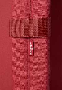 Červený látkový batoh s textúrovaným povrchom. Obsahuje tkaný popruh a malú bielu nálepku s červeným logom "Levi's".