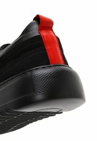 Chaussure noire en matériaux daim et cuir, avec une languette de talon en cuir rouge, une semelle noire texturée et un dessous blanc lisse.