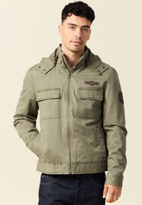 Man in olijfgroene zip-up jack met klepzakken en patches, staand met één hand in de zak tegen een neutrale achtergrond.