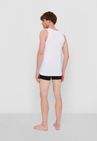 Vit bomullstopp utan ärmar med rund halsringning och vida armhål, i kombination med svarta shorts. Modellen är barfota och visar en avslappnad stil.