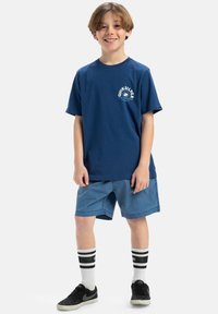 Námořnická modrá bavlněná tričko s logem Quiksilver, zkombinované se světle modrými šortkami. Nosí černé tenisky a bílé ponožky s černými pruhy.