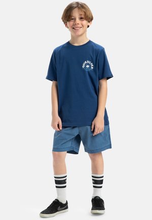 Marineblå bomulds T-shirt med Quiksilver-logo, parret med lyseblå shorts. Bærer sorte sneakers og hvide sokker med sorte striber.