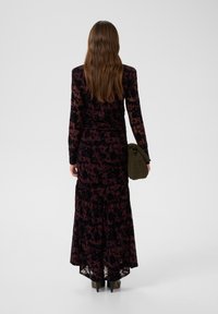 Langes lila Samtkleid mit Blumenmuster, angepasste Taille, lange Ärmel und ausgestellter Rock. Begleitet von einer grünen Clutch.