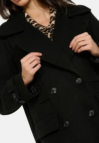 Schwarzer zweireihiger Wollmantel mit einer vorderen Klappe, großen Revers und vier Knöpfen. Darunter eine Bluse mit Leopardenmuster.