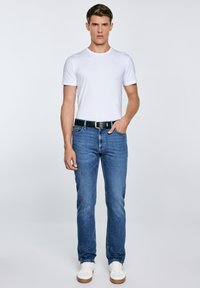 Férfi áll, fehér rövid ujjú pólót, kék farmert fekete övvel, fehér tornacipőt visel, egyik kezét a zsebében tartja, egyszínű háttér előtt.