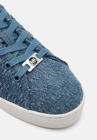 Bout de basket en denim bleu avec lacets assortis, semelle blanche et petite plaque argentée avec le logo MK sur les lacets.