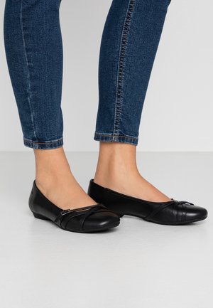 Stopy w czarnych skórzanych baletkach z krzyżującymi się paskami, zestawione z niebieskimi dżinsami typu skinny na jednolitym tle.