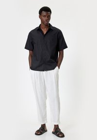 Camisa negra de manga corta con botones y un bolsillo en el pecho, combinada con pantalones de lino blancos y sandalias negras, de pie sobre un fondo neutro.