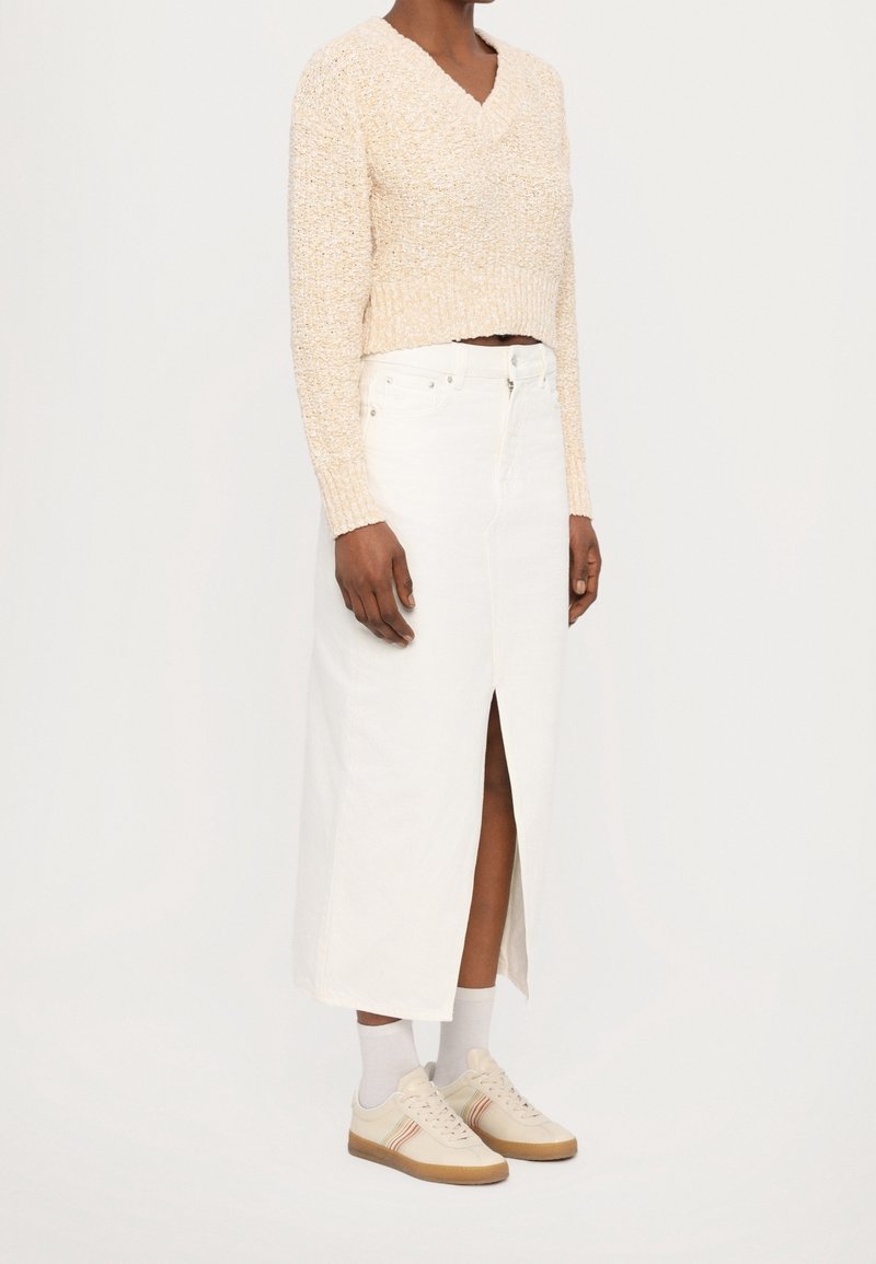 Personne portant un pull en tricot beige, une jupe midi blanche fendue sur le devant, des chaussettes blanches et des baskets beiges à semelles en gomme, vue de côté.