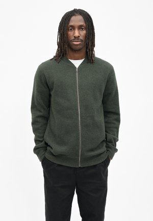 Man met dreadlocks tot op schouderlengte, gekleed in een donker groene trui met rits en een zwarte broek, staande met handen in de zakken tegen een witte achtergrond.