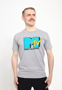 Šedé bavlněné tričko s velkým modrým logem "M" a žlutým logem "TV" s černým textem, umístěné na čistém bílém pozadí.