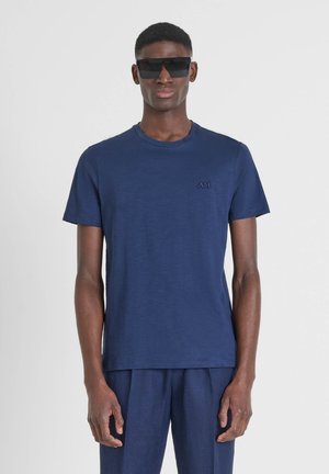 Camiseta de manga corta azul marino hecha de tejido suave, con cuello redondo y un logotipo sutil en el lado izquierdo del pecho. El modelo lleva unas grandes gafas de sol negras.