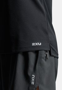 Sort atletisk trøje med struktureret stof; har en lille hvid "2XU" logo ved kanten. Kombineret med sorte shorts, også med "2XU" branding.