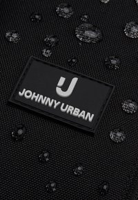 Čierna textúrovaná tkanina s kvapkami vody a čiernou obdĺžnikovou záplatou označenou "Johnny Urban" bielymi písmenami.
