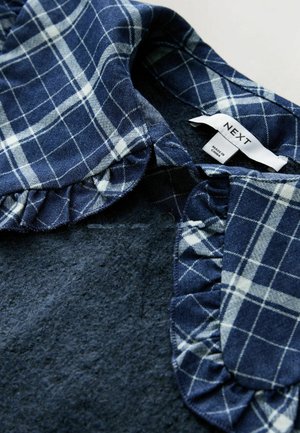 Guler în carouri bleumarin cu detaliu de volă pe un material texturat închis la culoare, inscripționat „NEXT”. Gulerul are linii albe în pattern de carouri.