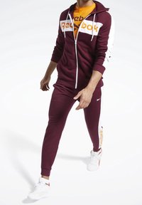Tuta color bordeaux con felpa con zip e logo bianco e arancione, abbinata a pantaloni coordinati e scarpe da ginnastica bianche.