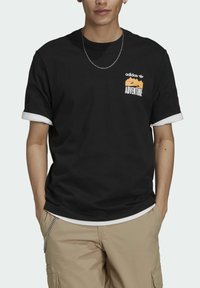 Czarny bawełniany t-shirt z białymi podwiniętymi rękawami, z pomarańczową i białą grafiką "ADVENTURE" oraz logo Adidas na klatce piersiowej.