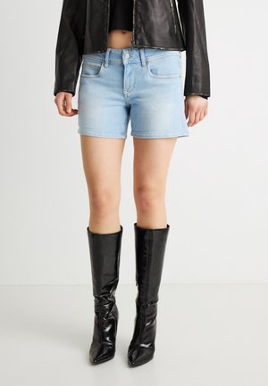 Persona con pantalones cortos de mezclilla azul claro, chaqueta de cuero negra recortada y botas negras de tacón hasta la rodilla sobre un fondo blanco liso.