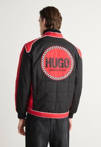 Czarna i czerwona pikowana kurtka z białymi akcentami, z dużym okrągłym logo "HUGO WINNING BLEND" na plecach i ściągaczami przy mankietach.