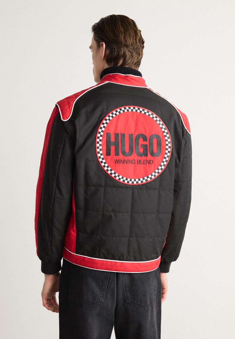 Czarna i czerwona pikowana kurtka z białymi akcentami, z dużym okrągłym logo "HUGO WINNING BLEND" na plecach i ściągaczami przy mankietach.