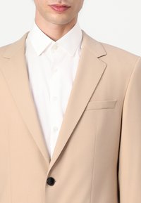 Blazer beige avec un bouton noir, poche poitrine simple et revers crantés, porté sur une chemise blanche à col. Tissu lisse et coupe ajustée.
