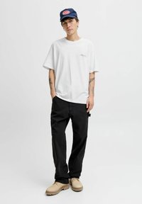 Baltas pārslu krekliņš ar lielu izmēru, melnas brīvas bikses, bēšas kurpes un tumši zilais cepure ar apaļu sarkanu logo. Vienkāršs, ikdienas dizains ar minimālistiskām detaļām.