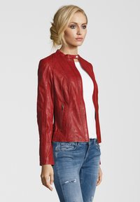 Rote Lederjacke mit hohem Kragen, Reißverschluss und zwei Seitentaschen. Über einem weißen Oberteil getragen und mit blauen, verwaschenen Jeans kombiniert.
