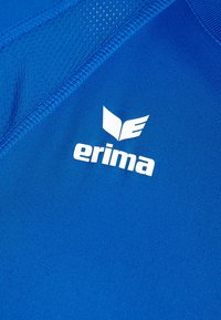 Primo piano di un tessuto sportivo blu con il logo bianco del marchio Erima e un dettaglio in rete traspirante vicino alla cucitura.