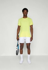 Kirkas keltainen urheiluhaitari, lyhyet hihat; yhdistetty valkoisiin shortseihin. Malli pitää mustaa ja sinistä tennismailaa, jalassa valkoiset kengät.