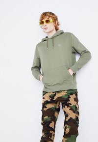Oliwkowy, damski sweatshirt z kapturem i przednią kieszenią, w połączeniu z brązowymi i zielonymi spodniami cargo w kamuflażu. Jasnożółte okulary przeciwsłoneczne.